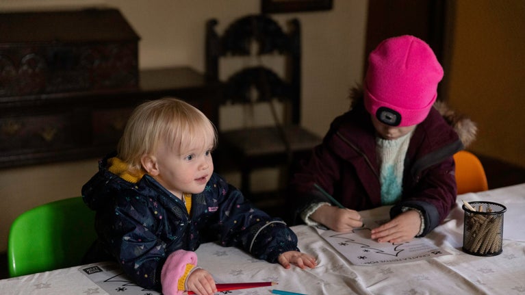 Two children sat at a table colouring. There is dark wooden furniture in the background.